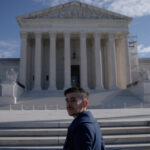 A man in a dark blue collared coat stands at the steps of the Supreme Court. He has brown hair and a moustache and is turning back to face the camera.