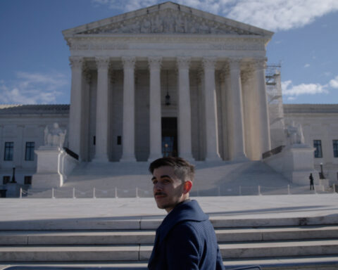 A man in a dark blue collared coat stands at the steps of the Supreme Court. He has brown hair and a moustache and is turning back to face the camera.