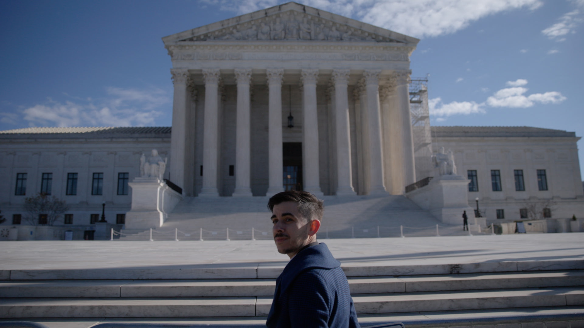A man in a dark blue collared coat stands at the steps of the Supreme Court. He has brown hair and a moustache and is turning back to face the camera.