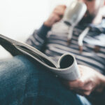 man reading magazine or newspaper and drinking coffee while relaxing on couch