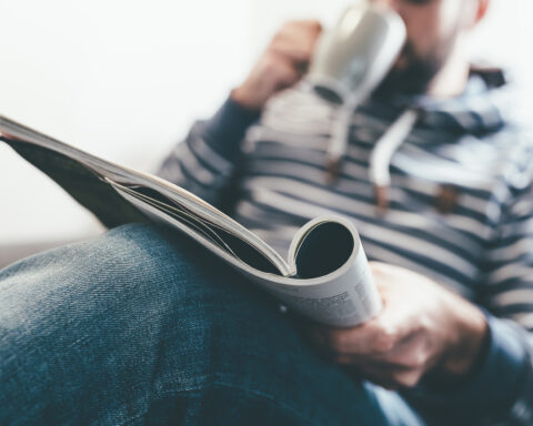 man reading magazine or newspaper and drinking coffee while relaxing on couch