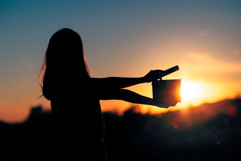 A woman holds a film clapper and is viewed in silhouette standing before a sunset.