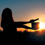 A woman holds a film clapper and is viewed in silhouette standing before a sunset.