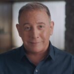 Actor Paul Reubens aka Pee Wee Herman sits in close up facing the camera. He is a 70-year old white man with greying hair, wearing a dark blue collared shirt.