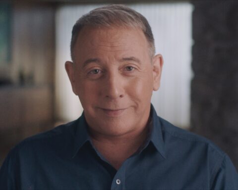 Actor Paul Reubens aka Pee Wee Herman sits in close up facing the camera. He is a 70-year old white man with greying hair, wearing a dark blue collared shirt.