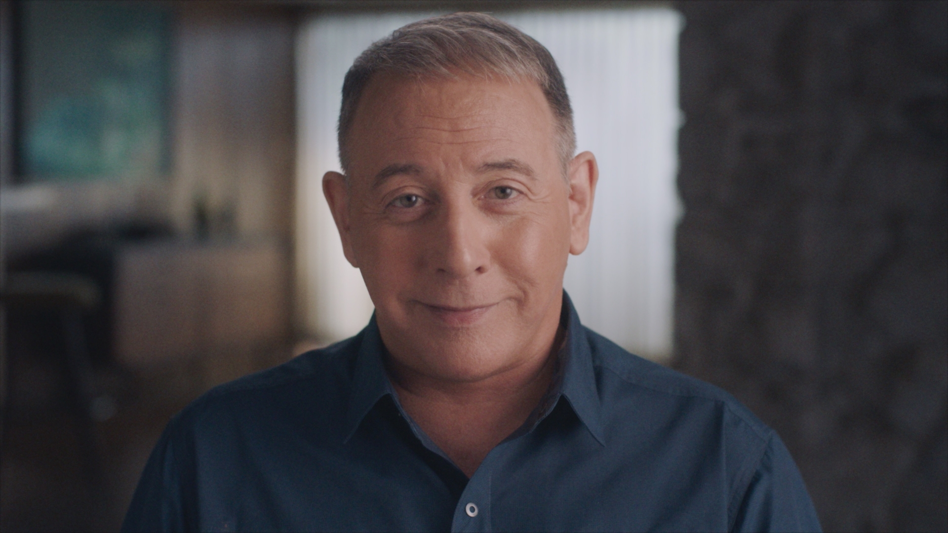 Actor Paul Reubens aka Pee Wee Herman sits in close up facing the camera. He is a 70-year old white man with greying hair, wearing a dark blue collared shirt.