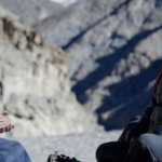 Two Nepali women sit on a mountainside. They are looking at the camera towards screen right and smiling. One slightly younger woman on the left is wearing a yellow puffy jacket and clasping her hands. The slightly older woman is on the right and is wearing a navy blue puffy jacket.