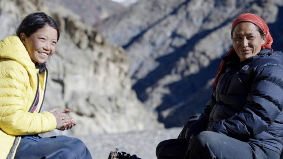 Two Nepali women sit on a mountainside. They are looking at the camera towards screen right and smiling. One slightly younger woman on the left is wearing a yellow puffy jacket and clasping her hands. The slightly older woman is on the right and is wearing a navy blue puffy jacket.