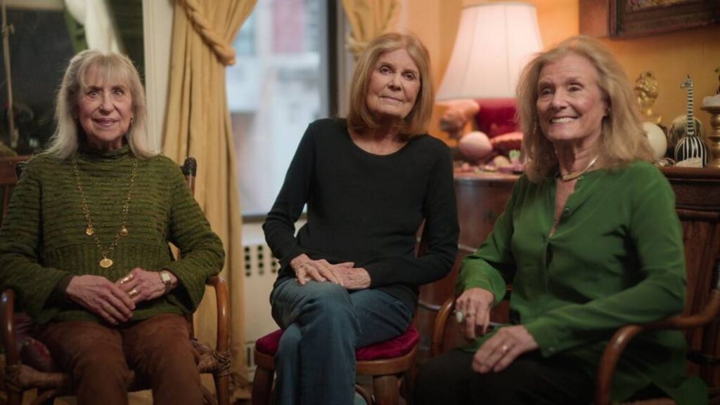 Suzanne Braun Levine (a woman with blond hair and wearing an olive green shirt), Gloria Steinem (with grey-brown hair and wearing a black sleeved shirt with blue jeans), and Letty Cottin Pogrebin (with blond hair, a dark green shirt, and black pants) are seated in a row facing the camera.