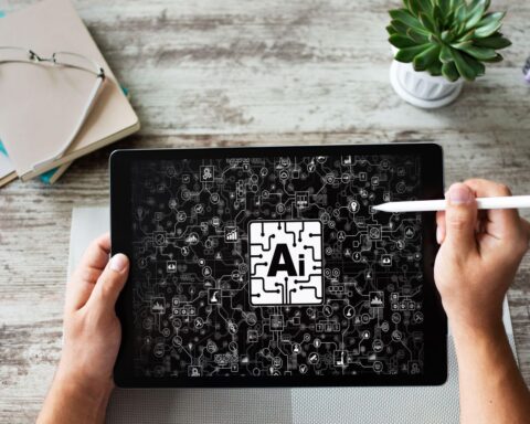 An image of a man's hand's holding a tablet and a wand above a messy desk. The tablet has a black screen with the letters AI in a white rectangle in the centre, while the screen is dotted with white lines and markings.