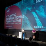 Two women stand on a stage. One is in the spotlight at a podium and the other is off to her side. There is a screen behind them advertising the Atlantic International Film Festival in red with white letters.