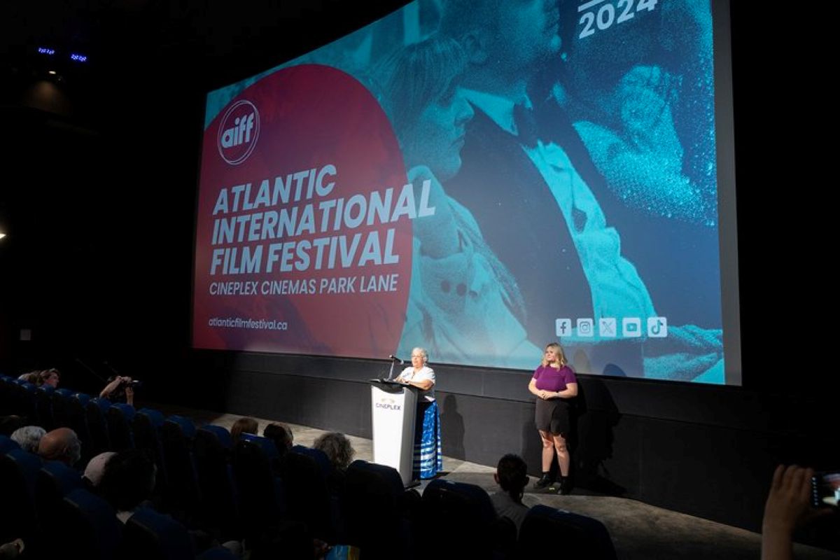 Two women stand on a stage. One is in the spotlight at a podium and the other is off to her side. There is a screen behind them advertising the Atlantic International Film Festival in red with white letters.