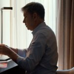A man sits at a desk with an open laptop. He is a white man with greying hair and is holding a paper letter. He is surrounded by windows with light filtering through the curtains.