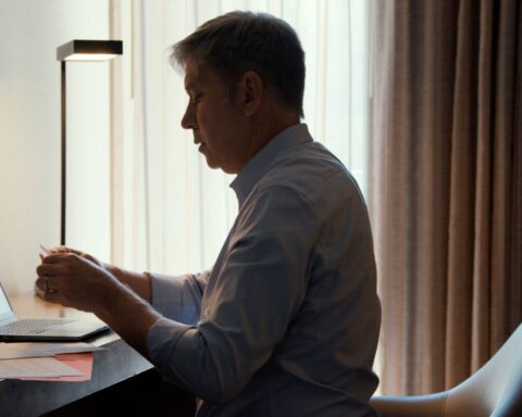 A man sits at a desk with an open laptop. He is a white man with greying hair and is holding a paper letter. He is surrounded by windows with light filtering through the curtains.