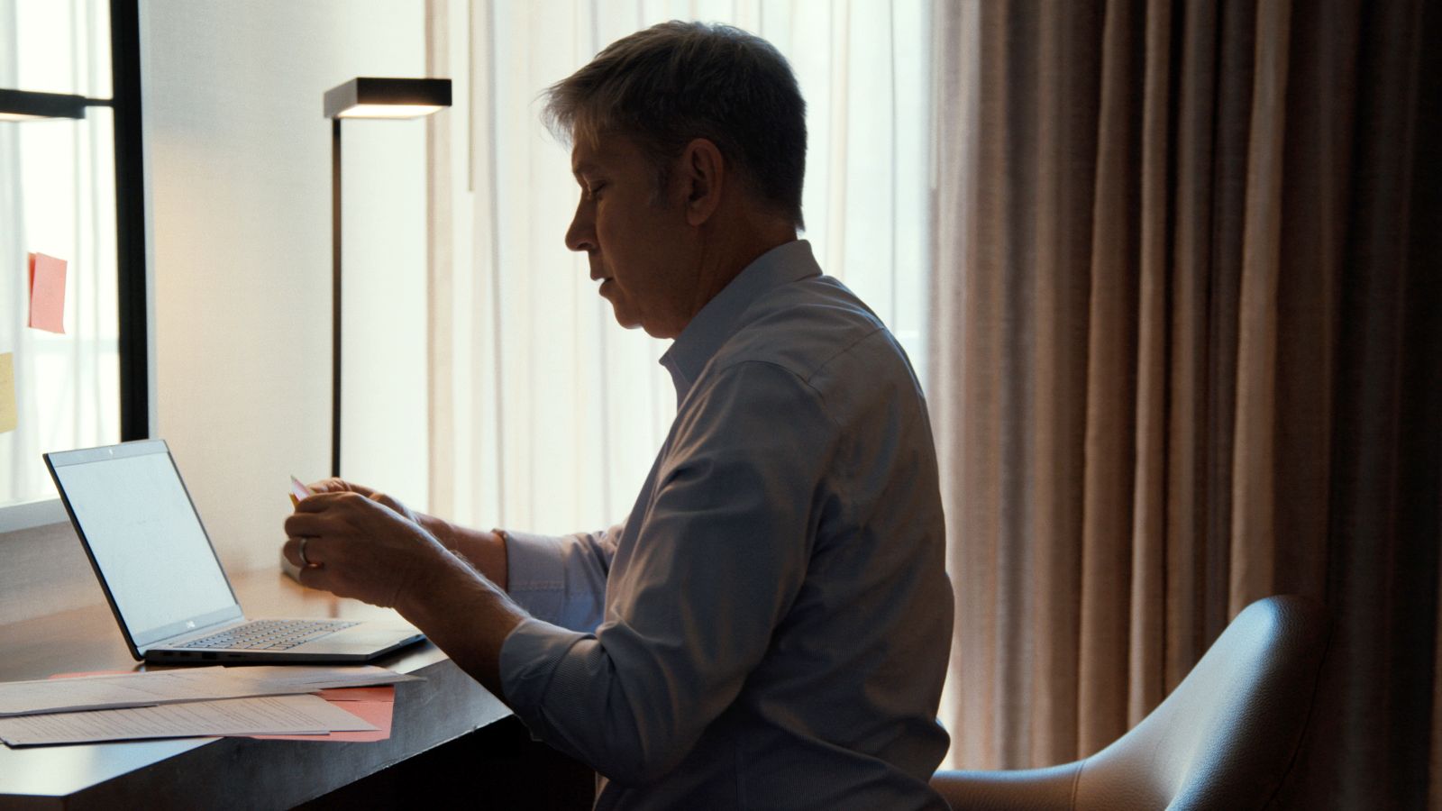 A man sits at a desk with an open laptop. He is a white man with greying hair and is holding a paper letter. He is surrounded by windows with light filtering through the curtains.