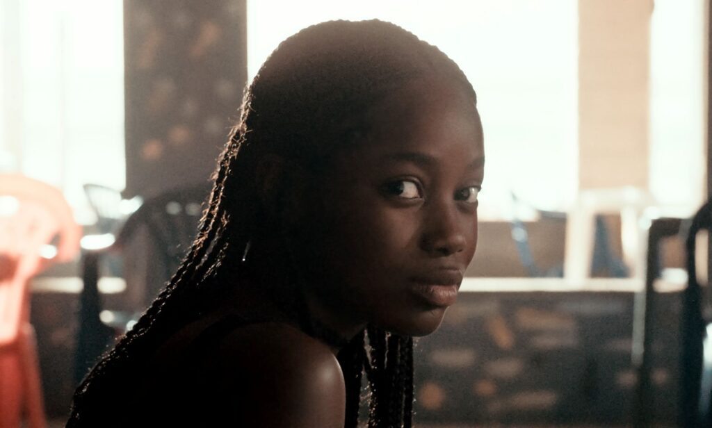A young Black womanwith braided hair is photographed in close-up. She looks directly at the camera with a playful, mysterious gaze.