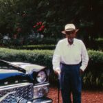 In a vintage photograph, an elderly Black man stands beside a dark blue car. The man is wearing a white shirt, blue slacks, and a white brimmed hat. He is holding a cane.