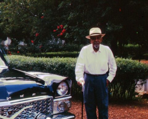 In a vintage photograph, an elderly Black man stands beside a dark blue car. The man is wearing a white shirt, blue slacks, and a white brimmed hat. He is holding a cane.