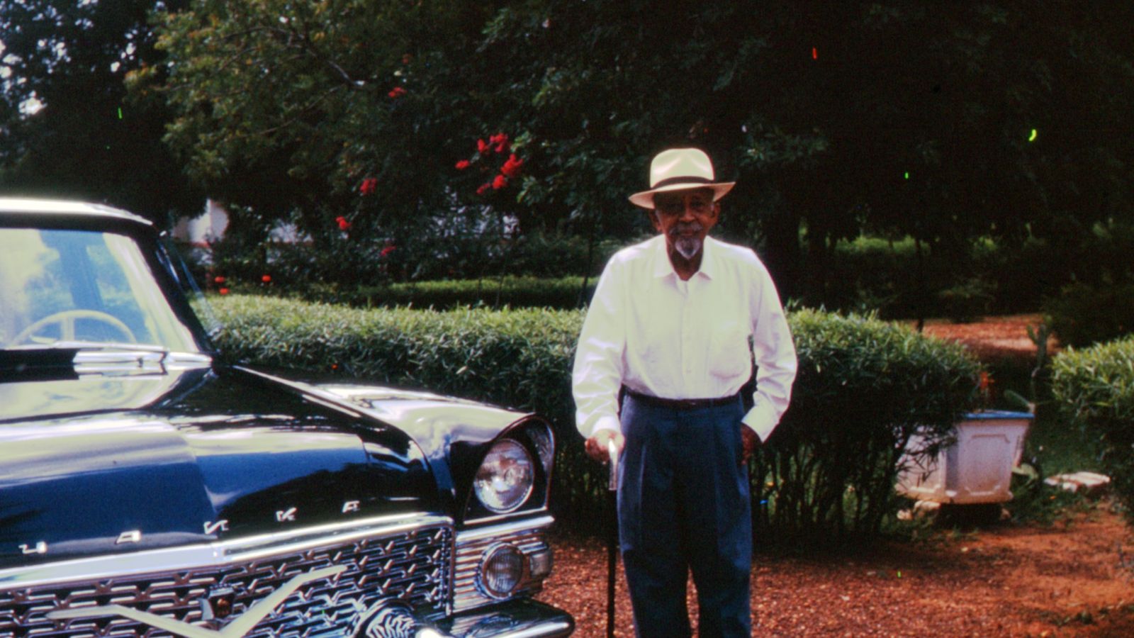 In a vintage photograph, an elderly Black man stands beside a dark blue car. The man is wearing a white shirt, blue slacks, and a white brimmed hat. He is holding a cane.