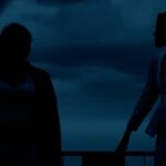 Two figures appear as black silhouettes against a darkened blue backdrop.