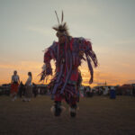 A man clad in cultural regalia dances with the sun setting in the backdrop