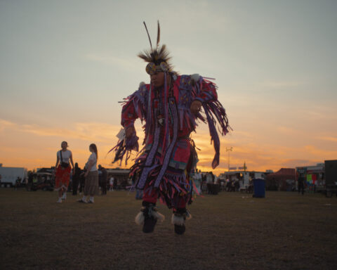 A man clad in cultural regalia dances with the sun setting in the backdrop