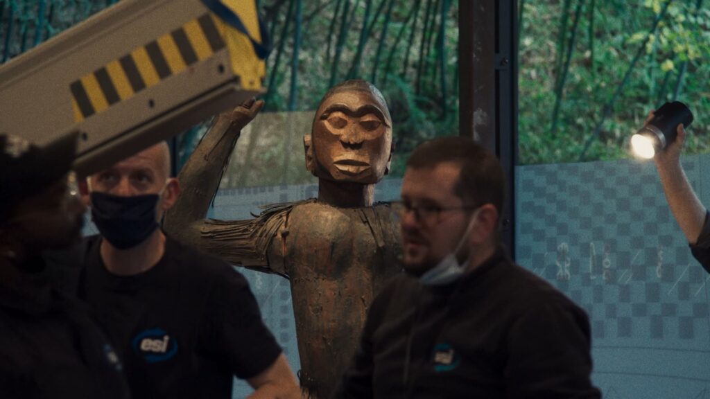 An African wooden statue with a raised hand is displayed in a museum. A white man with a COVID mask under his chin walks past it without looking.