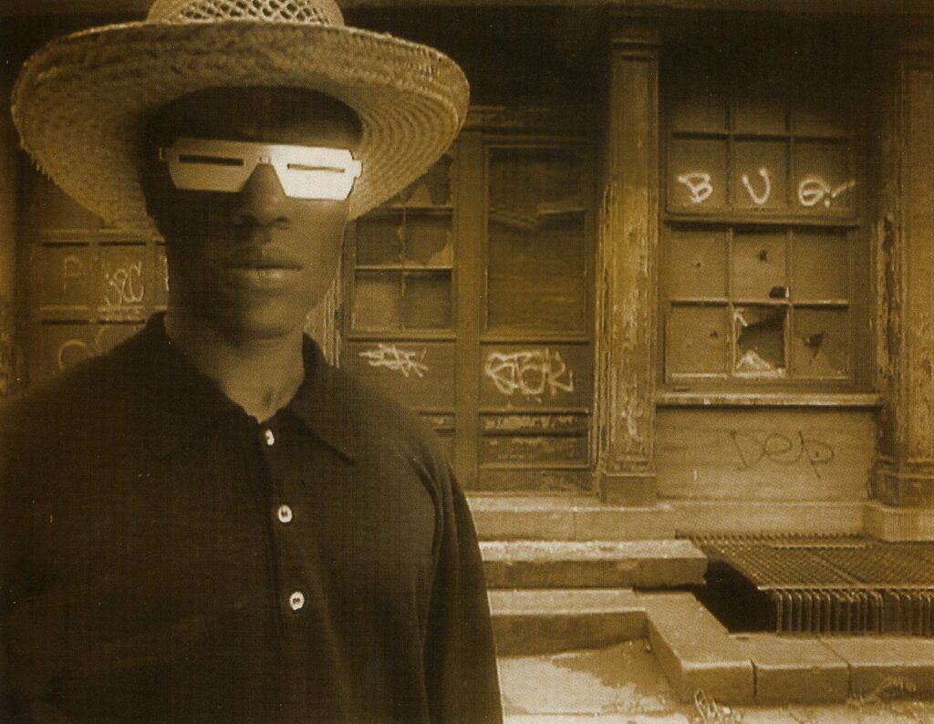 A Black man is photographed in medium shot and sepia tone. He is wearing solar protective glasses, a dark shirt, and a straw hat.