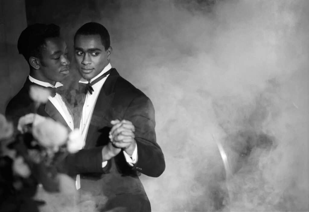 Two Black men in tuxedos dance closely together. They are photographed in black and white and the image that surrounds them has a smoky haze.