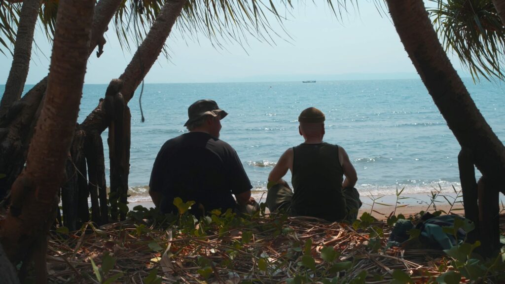 A view from behind two people as they sit on the beach and look out at the ocean. They are framed by palm trees.