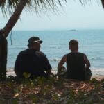 A view from behind two people as they sit on the beach and look out at the ocean. They are framed by palm trees.