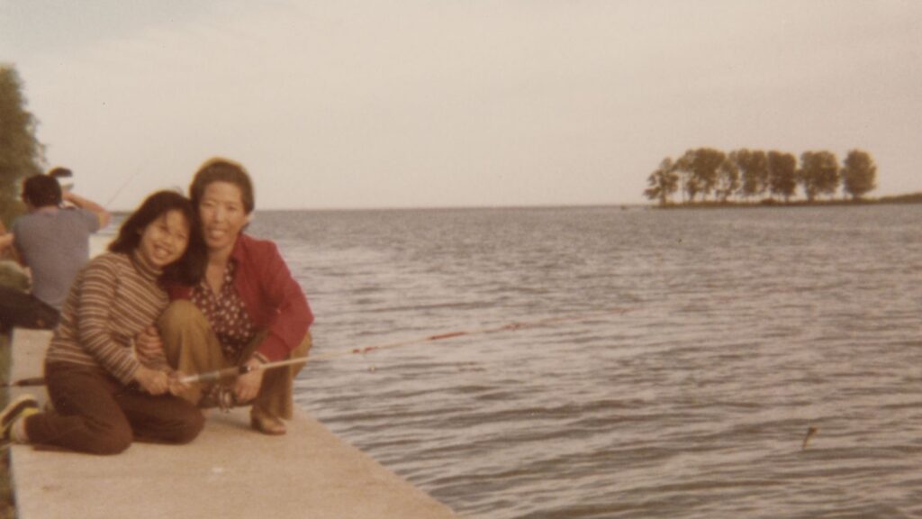 An old photograph of a Korean mother and teen daughter, kneeling together by the water. They are smiling.