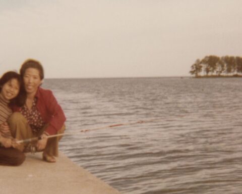 An old photograph of a Korean mother and teen daughter, kneeling together by the water. They are smiling.