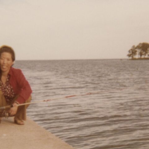 An old photograph of a Korean mother and teen daughter, kneeling together by the water. They are smiling.