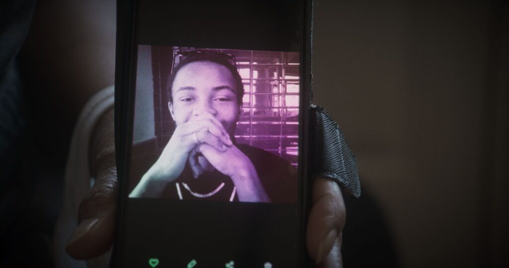 A young Black man is pictured on a phone screen. He has his hands clasped in front of his face and appears to be smiling.