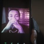 A young Black man is pictured on a phone screen. He has his hands clasped in front of his face and appears to be smiling.