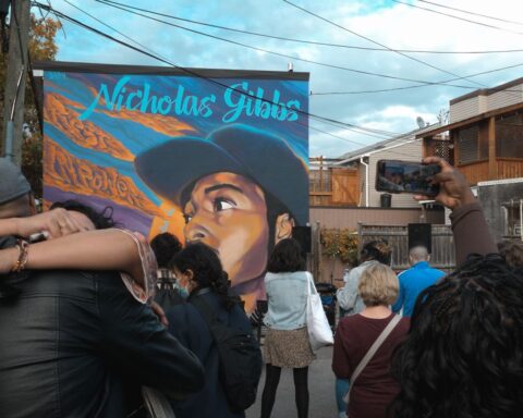 Two people hug amid a rally in the streets of Montreal. Behind them is a street art portrait of Nicholas Gibbs, a 23-year-old Black man.