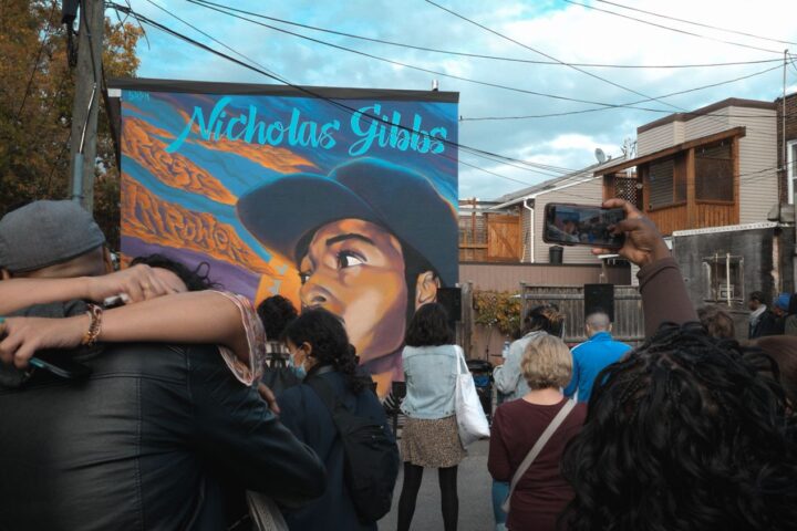 Two people hug amid a rally in the streets of Montreal. Behind them is a street art portrait of Nicholas Gibbs, a 23-year-old Black man.