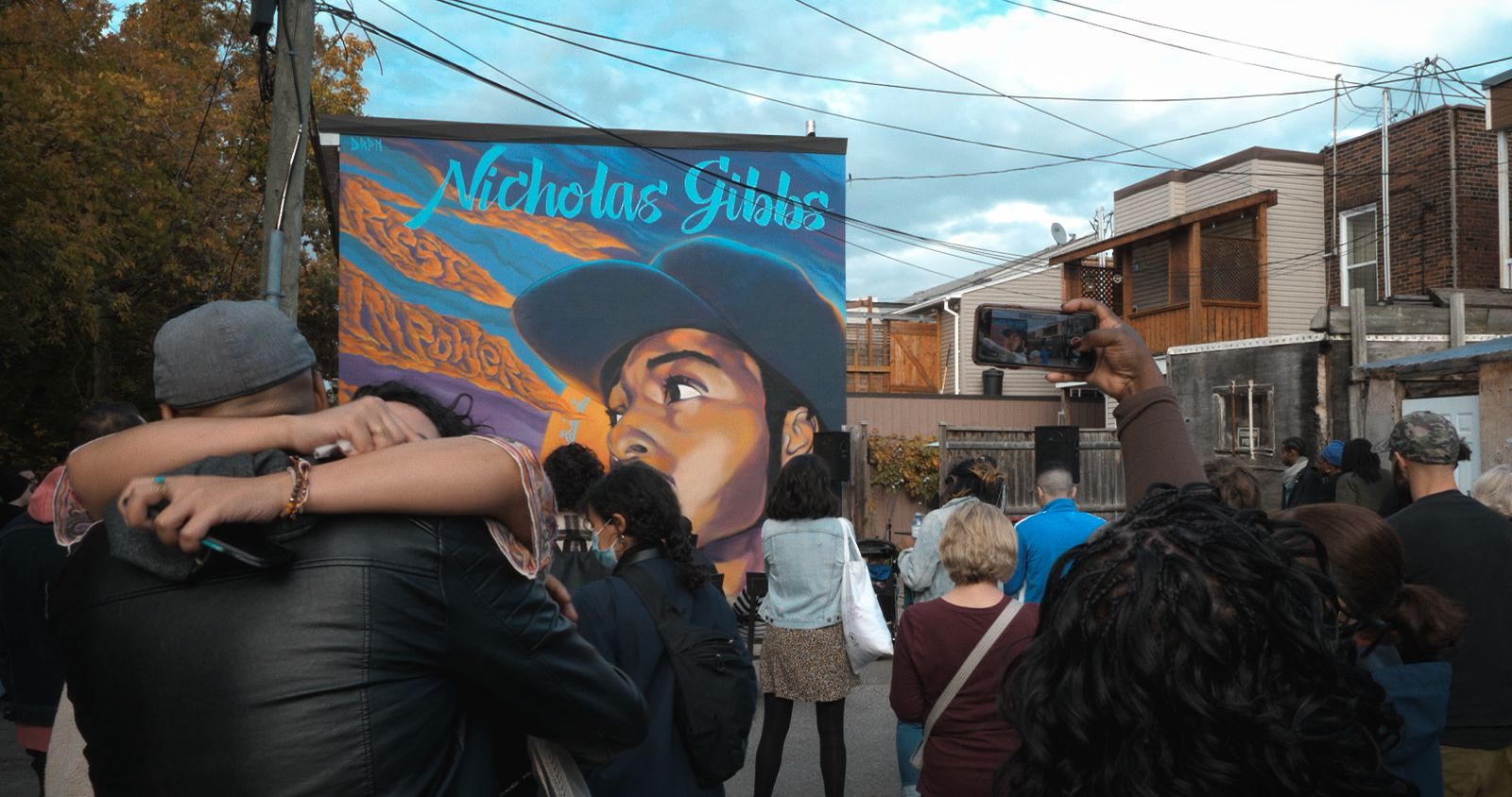 Two people hug amid a rally in the streets of Montreal. Behind them is a street art portrait of Nicholas Gibbs, a 23-year-old Black man.