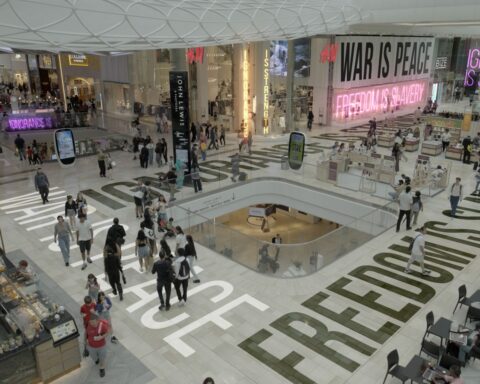 A crowded shopping mall contains people walking atop superimposed text of words including freedom and peace.