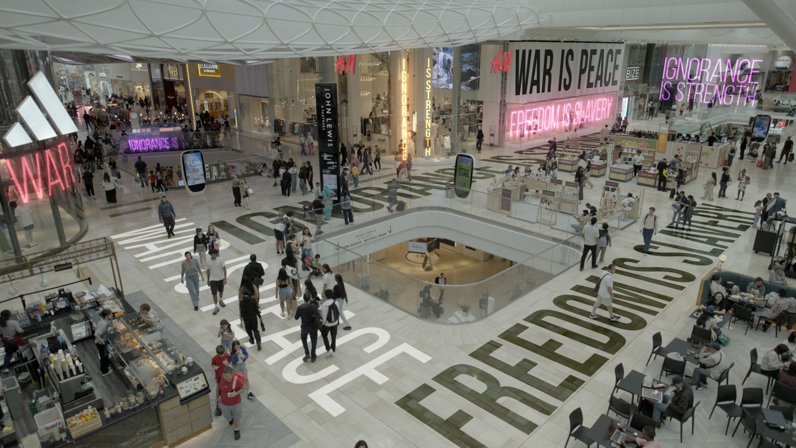 A crowded shopping mall contains people walking atop superimposed text of words including freedom and peace.