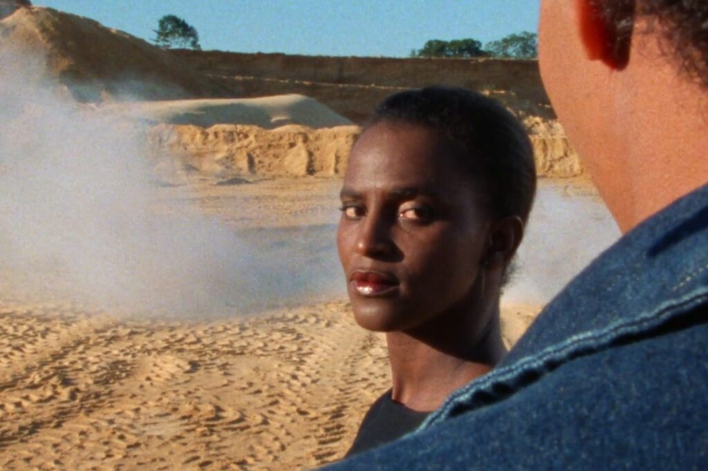 A young Black woman looks at the camera and over the shoulder of a white man, who is standing directly in front of the camera and viewed from behind.