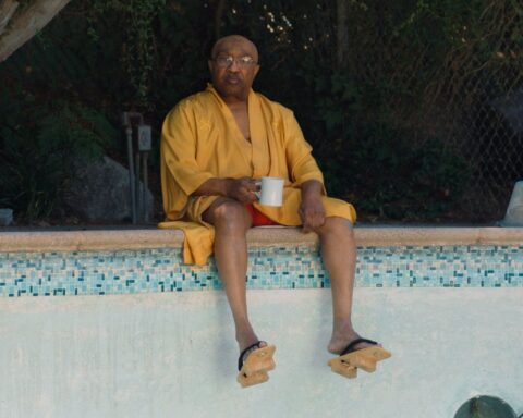 An older Black man in an orange robe sits on the edge of an empty swimming pool. He is holding a cup of coffee and his feet are dangling over the edge of the pool.