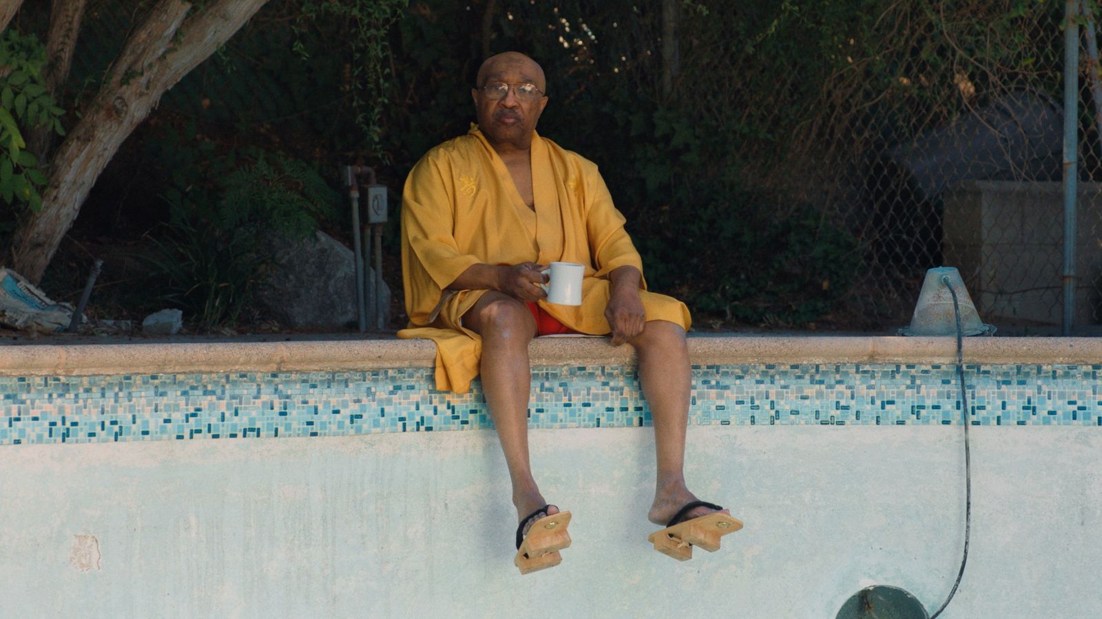 An older Black man in an orange robe sits on the edge of an empty swimming pool. He is holding a cup of coffee and his feet are dangling over the edge of the pool.