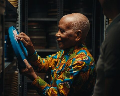 93-year-old Ghanaian filmmaker Chris Hesse holds up a reel of film and is pointing at the label and smiling. He has grey hair and is wearing a multicolour patterned shirt.