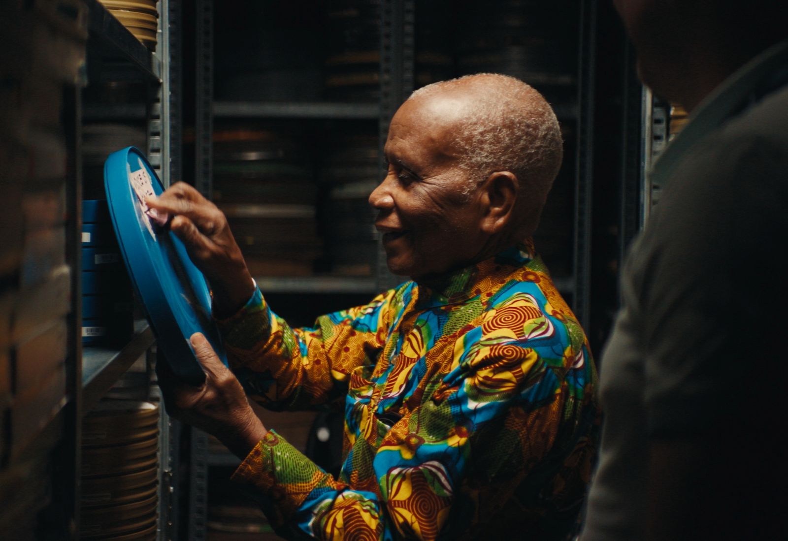 93-year-old Ghanaian filmmaker Chris Hesse holds up a reel of film and is pointing at the label and smiling. He has grey hair and is wearing a multicolour patterned shirt.