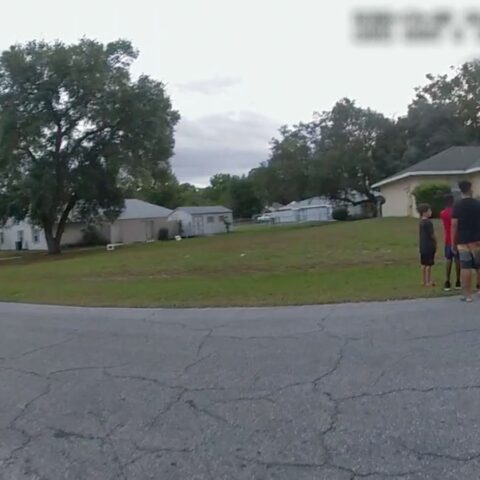A image from a police body camera shows a neighbourhood and a field. There is a police car on the right with a crowd of people nearby.