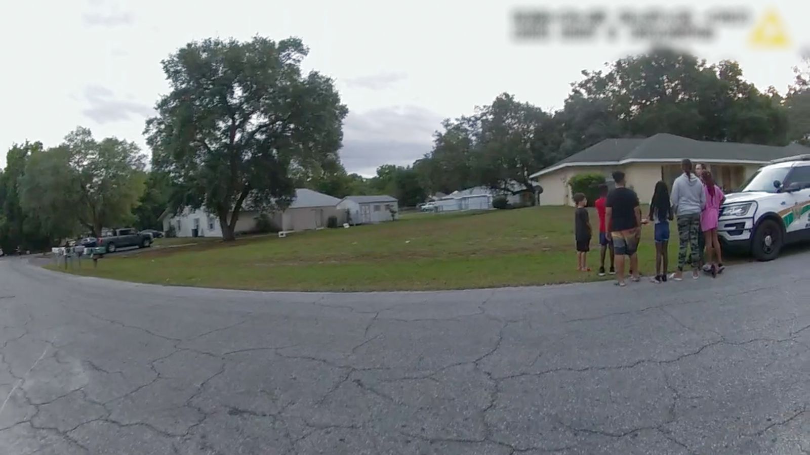 A image from a police body camera shows a neighbourhood and a field. There is a police car on the right with a crowd of people nearby.