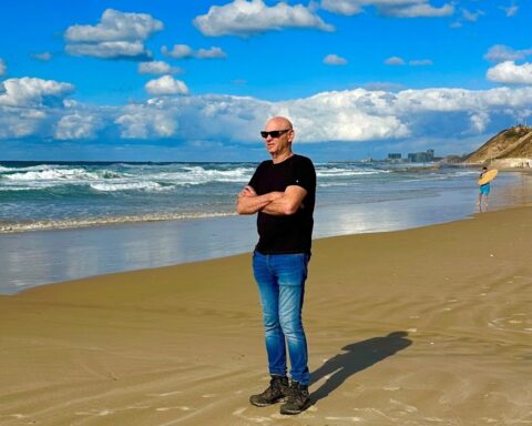 A man in a black t-shirt and blue jeans stands on a beach. His arms are crossed and he is looking out at the water.