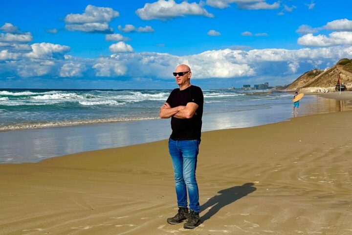 A man in a black t-shirt and blue jeans stands on a beach. His arms are crossed and he is looking out at the water.